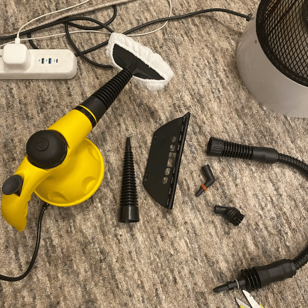 Yellow handheld steam cleaner with multiple nozzle attachments laid out on a carpeted floor